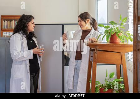 Zwei junge Wissenschaftlerinnen in weißen Laborkitteln, die während einer Pause plaudern, eine hält eine Tasse und die andere ein Glas Wasser, stehend in der Nähe eines Holztisches Stockfoto