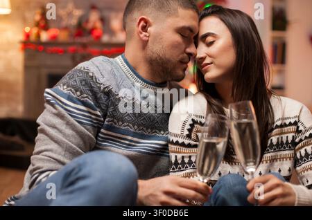 Ein verliebtes Paar schließt die Augen am Weihnachtstag, während es Champagnergläser hält, und teilt einen zarten festlichen Moment zu Hause mit warmer saisonaler Intimität. Stockfoto