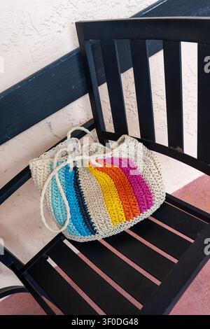 eine beige Häkel-Strand-Schultertasche mit lebendigen mehrfarbigen Streifen, die auf einem schwarzen Stuhl ruht und Sommer-Mode-Stil präsentiert Stockfoto