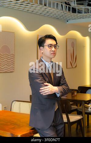 Selbstbewusster asiatischer Geschäftsmann in einem hochwertig maßgeschneiderten braunen Anzug, der elegant in einer modernen Indoor-Café-Lounge posiert, attraktives männliches Modelporträt. Stockfoto