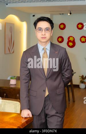 Selbstbewusster asiatischer Geschäftsmann in einem hochwertig maßgeschneiderten braunen Anzug, der elegant in einer modernen Indoor-Café-Lounge posiert, attraktives männliches Modelporträt. Stockfoto
