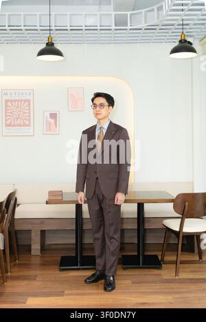 Selbstbewusster asiatischer Geschäftsmann in einem hochwertig maßgeschneiderten braunen Anzug, der elegant in einer modernen Indoor-Café-Lounge posiert, attraktives männliches Modelporträt. Stockfoto