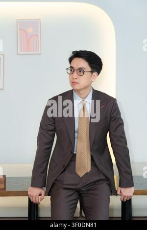 Selbstbewusster asiatischer Geschäftsmann in einem hochwertig maßgeschneiderten braunen Anzug, der elegant in einer modernen Indoor-Café-Lounge posiert, attraktives männliches Modelporträt. Stockfoto