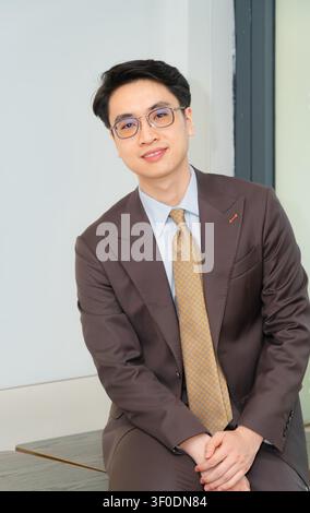 Selbstbewusster asiatischer Geschäftsmann in einem hochwertig maßgeschneiderten braunen Anzug, der elegant in einer modernen Indoor-Café-Lounge posiert, attraktives männliches Modelporträt. Stockfoto