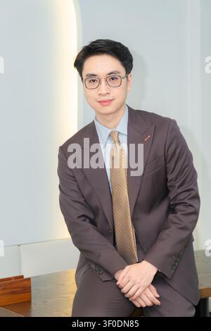 Selbstbewusster asiatischer Geschäftsmann in einem hochwertig maßgeschneiderten braunen Anzug, der elegant in einer modernen Indoor-Café-Lounge posiert, attraktives männliches Modelporträt. Stockfoto