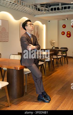 Selbstbewusster asiatischer Geschäftsmann in einem hochwertig maßgeschneiderten braunen Anzug, der elegant in einer modernen Indoor-Café-Lounge posiert, attraktives männliches Modelporträt. Stockfoto