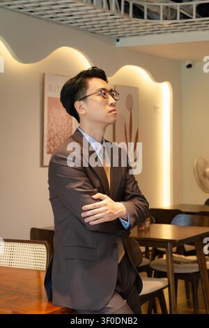 Selbstbewusster asiatischer Geschäftsmann in einem hochwertig maßgeschneiderten braunen Anzug, der elegant in einer modernen Indoor-Café-Lounge posiert, attraktives männliches Modelporträt. Stockfoto