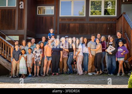 Große Gruppe von Verwandten und Freunden verschiedenen Alters, die zusammen auf den vorderen Stufen eines braunen Holzhauses stehen und an einem hellen Tag in legerer Kleidung lächeln Stockfoto