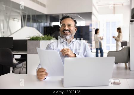 Zufriedener Geschäftsmann sitzt am Schreibtisch mit offenem Laptop und Papieren Stockfoto