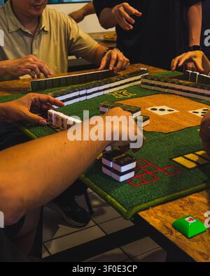 Ein ungeposiertes, nah aufgenommenes Foto von Spielern, die während eines lebhaften Mahjong-Spiels an einem Holztisch Spielsteine auf einer grünen Filzunterlage organisieren und bewegen. Stockfoto