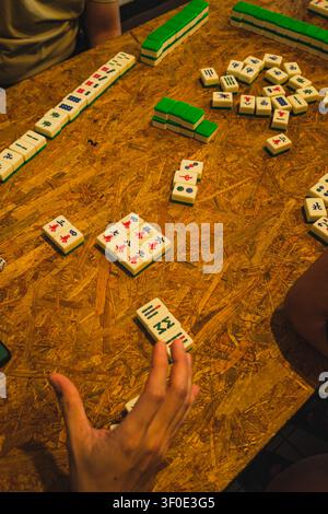 Eine Nahaufnahme einer Hand eines Spielers, die einen verstreuten Stapel Mahjong-Steine auf einem Tisch aus Holzpartikelbrett handhabt, wobei einige Steine mit der Bildseite nach oben liegen und Char zeigen Stockfoto