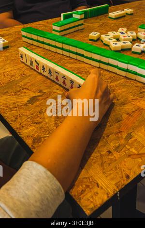 Eine Hochwinkel-Aufnahme, Nahaufnahme einer Hand eines Spielers, der weiße Mahjong-Steine aus Harz mit grünen Rückseiten auf einem Tisch aus Holzspanplatten ordnet, mit Stapel Stockfoto