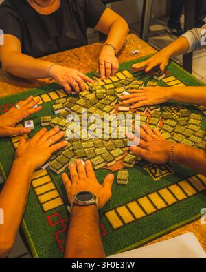 Eine Hochwinkel-, Nahaufnahme mehrerer Spielerhände, die glitzernde Mahjong-Steine mit grünem Rücken verdeckt auf einem grünen, strukturierten Tisch mischen und schieben Stockfoto