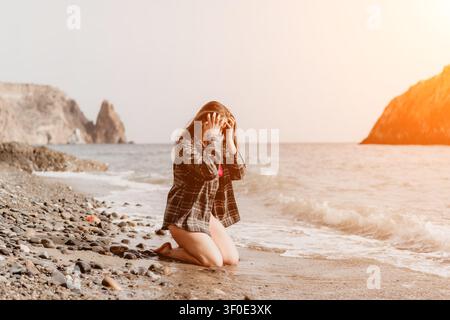 Pebble Beach Frau kniet am Ufer mit den Händen am Kopf in Not, während Wellen gegen felsige Klippen während des Sonnenuntergangs schlagen Stockfoto