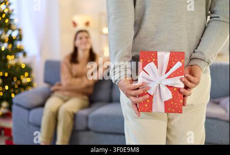Weihnachten zu zweit, Mann hält eine Geschenkschachtel, um sie der auf dem Sofa sitzenden Frau zu geben Stockfoto