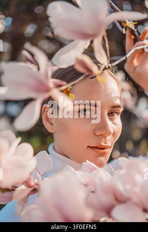 Porträt einer jungen Frau mit Magnolienblüte, die durch rosa Frühlingsblumen in einem sonnigen Gartenpark blickt Stockfoto