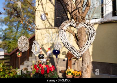 Rustikales Weidenherz hängt von Baumästen in einem sonnigen Hof neben bunten Blumen und einer gemütlichen Hausfassade. Handgemachte Ornamente, weicher Bokeh und warm Stockfoto