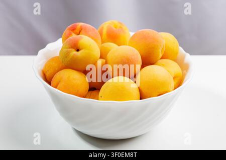 Es gibt ein Dutzend frische Aprikosen in einer tiefen weißen Schale. Die Früchte haben unterschiedliche Schattierungen – von hellgelb bis sattem Orange. Die Haut ist glänzend, slig Stockfoto