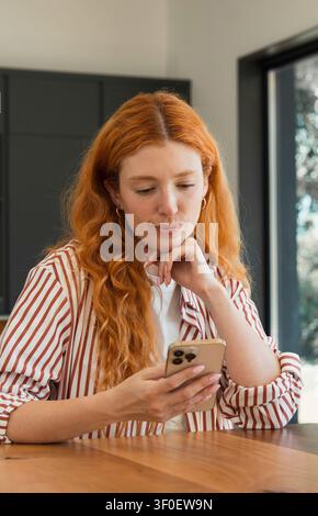 Eine rothaarige Frau sitzt an einem Holztisch im natürlichen Licht und konzentriert sich auf ihr Smartphone. Lässiges Indoor-Porträt, das Konzentration und modernen Lebensstil vermittelt Stockfoto