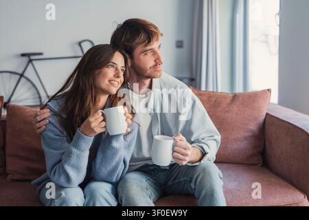 Glückliches, liebevolles Paar, das sich auf der Couch entspannt, heiße Getränke, Tee oder Kaffee trinkt, weiße Tassen hält, lächelnder junger Mann und Frau plaudern, reden, verbringen Stockfoto