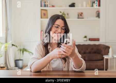 Eine lächelnde Frau sitzt an einem Holztisch in einem gemütlichen Wohnzimmer und nutzt glücklich ihr Smartphone, um zu texten, in sozialen Medien zu surfen und mit Freunden in Kontakt zu bleiben, während Stockfoto