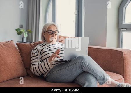 Seniorin mit Brille und gestreiftem Pullover sitzt bequem auf einem Sofa und nutzt einen Laptop zu Hause, multitaskt zwischen Remote-Arbeit, Surfen und s Stockfoto