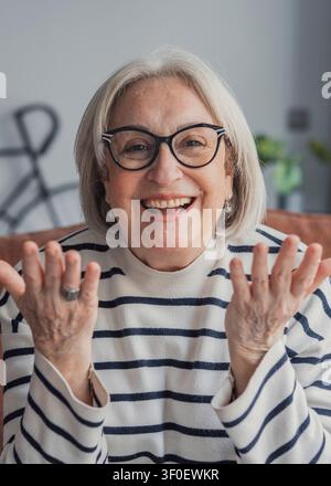Lächelnde ältere Frau mit Brille lacht herzlich in einer gemütlichen Wohnzimmerszene. Lässiger gestreifter Pullover und offene Hände vermitteln Freude, Freundlichkeit und com Stockfoto