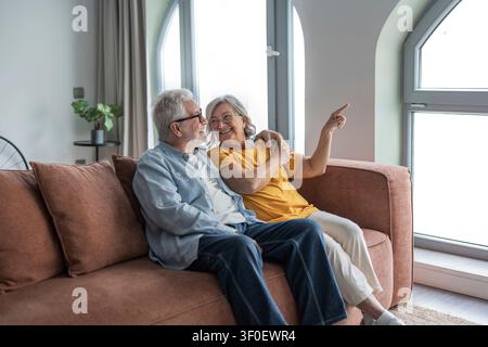 Ein glückliches Seniorenpaar sitzt eng beieinander auf einem Sofa in einem hellen Wohnzimmer, lächelt und zeigt auf das Fenster, während sie einen warmen, liebevollen Moment teilen Stockfoto