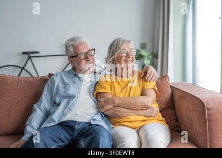 Ein älteres Ehepaar sitzt zusammen auf einem bequemen Sofa in ihrem Wohnzimmer, lächelt und teilt einen ruhigen, liebevollen Moment. Warmes natürliches Licht Stockfoto