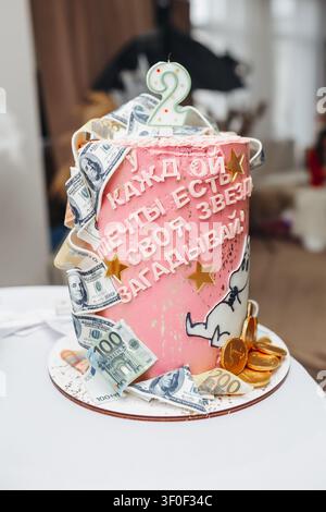 Rosa Kuchen, verziert mit Geldscheinen und einer Kerze. Goldmünzen und Geldscheine fließen an seinen Seiten herab. Geburtstagsnachricht in verspielter kyrillischer Schrift. Stockfoto