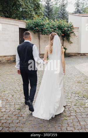 Braut und Bräutigam gehen Hand in Hand, mit dem Rücken zur Kamera. Weiches Licht unterstreicht ihre stille Freude. Ein gepflasterter Weg führt durch Grün und Steinmauern. Romantisch Stockfoto