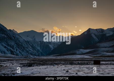 Blick auf das Khunjerab-Checkpost-Gebiet bei Sonnenuntergang mit schneebedeckten Karakorum-Berggipfeln, felsigem Gelände und goldenem Licht auf den Wolken in Hunza, Gilgit Stockfoto