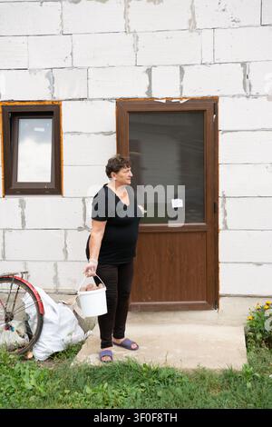 Dies ist eine Außenaufnahme einer Frau, die einen Eimer mit Inhalt vor ihrem Backsteingebäude hält. Ihr Haus hat Fenster und eine Tür. Ein Fahrrad Stockfoto