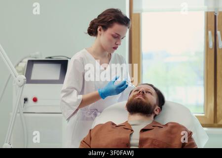 Junge erwachsene kaukasische Frau, Klinikerin, bereitet eine Gesichtsinsjektion für einen mittelalten kaukasischen Mann in einer ästhetischen Klinik vor. Ideal für Hautpflegebehandlungen, Marketing für kosmetische Verfahren Stockfoto