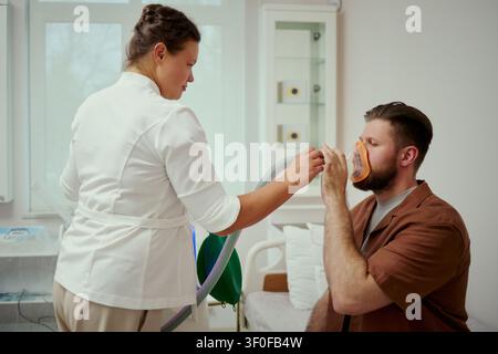Kaukasische Ärztin mittleren Alters unterstützt einen kaukasischen jungen erwachsenen Mann, der während eines respiratorischen Tests in der Klinik eine Sauerstoffmaske verwendet. Nützlich für Gesundheitswesen, Lungenpflege, Diagnosemarketing Stockfoto