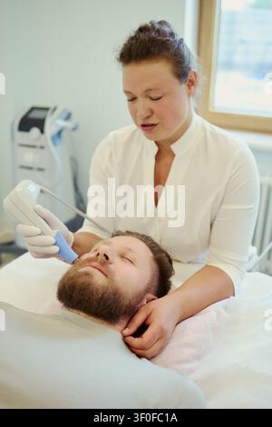 Vertikale Aufnahme einer mittelalten kaukasischen Frau, die während der Klinikbehandlung eine Ultraschalltherapie an einem mittelalten kaukasischen Mann durchführt. Ideal für Gesundheitswesen, Rehabilitation, Diagnostik Stockfoto