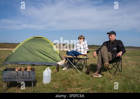 Paar entspannt in Campingstühlen neben einem grünen Zelt, grillt auf einem Mangal gespießtes Fleisch und teilt eine sonnige Mahlzeit im Freien auf einer Wiese während der Freizeit Stockfoto