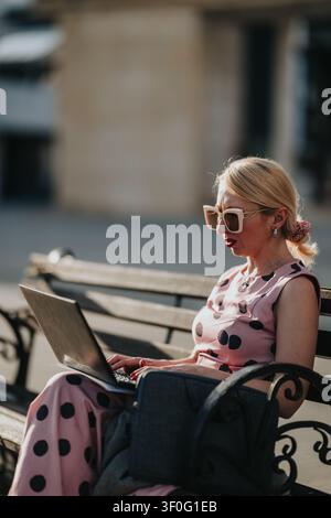 Frau arbeitet an einem Laptop im Freien auf einer Bank in einem rosa gepunkteten Kleid Stockfoto