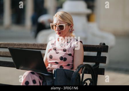 Reife Frau arbeitet an einem Laptop im Freien in einem rosa gepunkteten Kleid und einer Sonnenbrille auf einer Bank Stockfoto