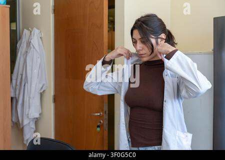 Junge Wissenschaftlerin, die in einem hellen Laborraum einen weißen Laborkittel anpasst und anzieht, wobei Laborkittel an einem Türhaken im Hintergrund sichtbar hängen Stockfoto
