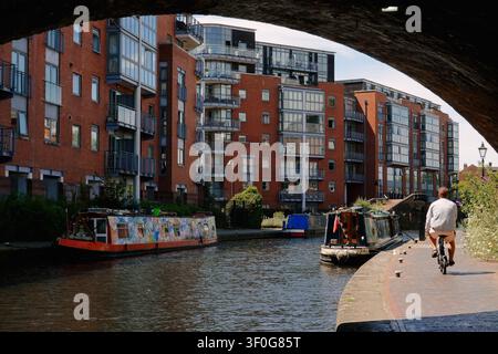 Ein Radfahrer fährt entlang des Kanaluferweges unter einer Brücke vorbei an modernen Wohnungen und schmalen Booten in Birmingham, UK Stockfoto