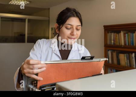 Junge Wissenschaftlerin in weißem Laborkittel lächelt, während sie einen orangefarbenen Ordner aus einem Aktenschrank zieht, mit einem Bücherregal farbenfroher Bücher, das sanft verschwommen ist Stockfoto