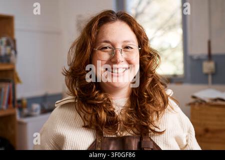 Lächelnde rothaarige Töpferwerkstatt-Besitzerin mit Schürze und Brille in kreativem Kleinunternehmens-Workshop Stockfoto