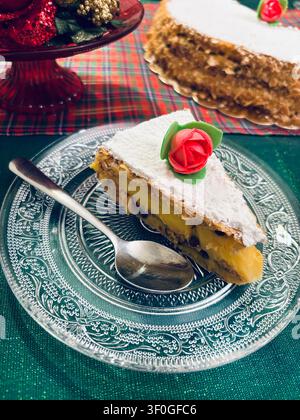 Köstliche Tortenstück, mit Puderzucker bestreut, verziert mit einer roten Blume auf einem Vintage-Glasplatte. Festlicher Weihnachts-Hintergrund mit karierter Tischdecke. Stockfoto