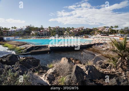 Urlauber entspannen sich auf Liegestühlen unter orangefarbenen Sonnenschirmen rund um ein großes Meerwasserschwimmbad neben dem Atlantischen Ozean in einem Küstenort in Costa Adeje an der Südküste von Teneriffa auf den Kanarischen Inseln, Spanien. Fotografiert am 18. Januar 2026. Stockfoto