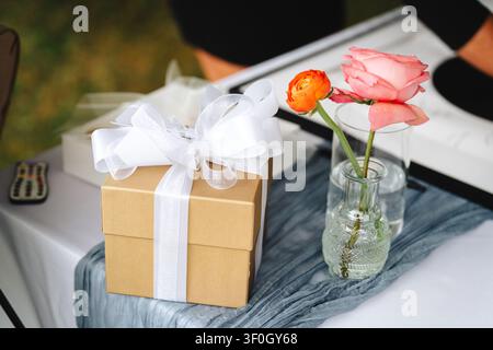 Verpackte Geschenkschachtel mit weißer Schleife und Rosen auf dem Tisch für Hochzeitsgäste Stockfoto