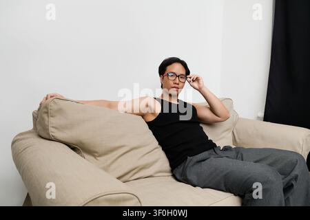 Ein stilvoller junger Mann sitzt lässig auf einer bequemen Couch und strahlt Selbstbewusstsein in seinem eleganten Outfit aus. Stockfoto