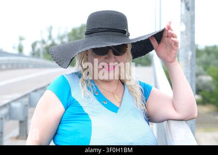 Selbstbewusste lächelnde reife Frau über 50, die einen breiten schwarzen Sonnenhut anpasst, während sie an einem warmen Sommertag auf einer Fußgängerbrücke steht. Aktiver Lebensstil Stockfoto