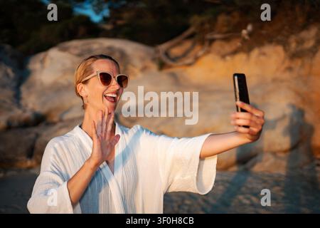 Glückliche Frau, die während eines Videoanrufs am Strand bei Sonnenuntergang winkt Stockfoto