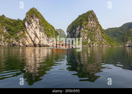 Große Kalkstein-Karstformationen und ein reflektierendes traditionelles hölzernes Touristenjunkboot in der Halong-Bucht, Vietnam Stockfoto